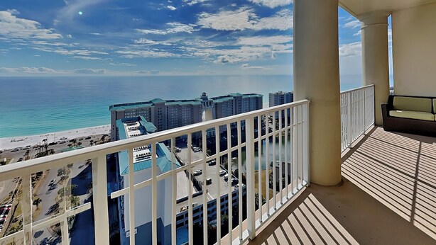 Un balcón con vistas a la playa y al océano, barandillas blancas y un edificio con tejados azules al fondo.