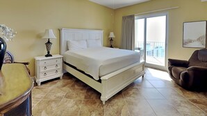 Habitación de hotel con una cama grande, ventilador de techo, mesita de noche con lámparas, una silla y vistas al balcón exterior.