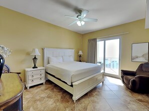 A hotel room with a large bed, a ceiling fan, a nightstand with lamps, a chair, and a view of the outside balcony. - Ariel Dunes Penthouse #2406 | Luxe Penthouse, Huge Wraparound Balcony (Miramar Beach)