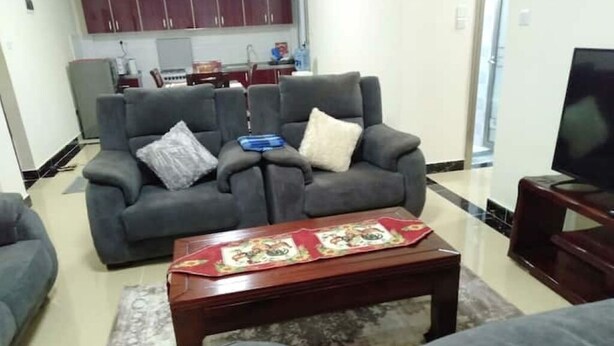 A living room with a grey sofa set, a wooden coffee table with a decorative runner, and a television on a stand. - Stylish 2BR Kilimani Retreat | Lift, Fast WiFi & Family-Friendly Comfort (Nairobi)