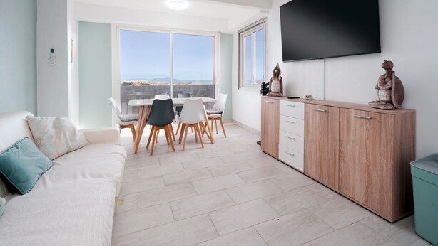 Una sala de estar moderna con un sofá, un comedor con sillas, un televisor montado en la pared y vistas al exterior.