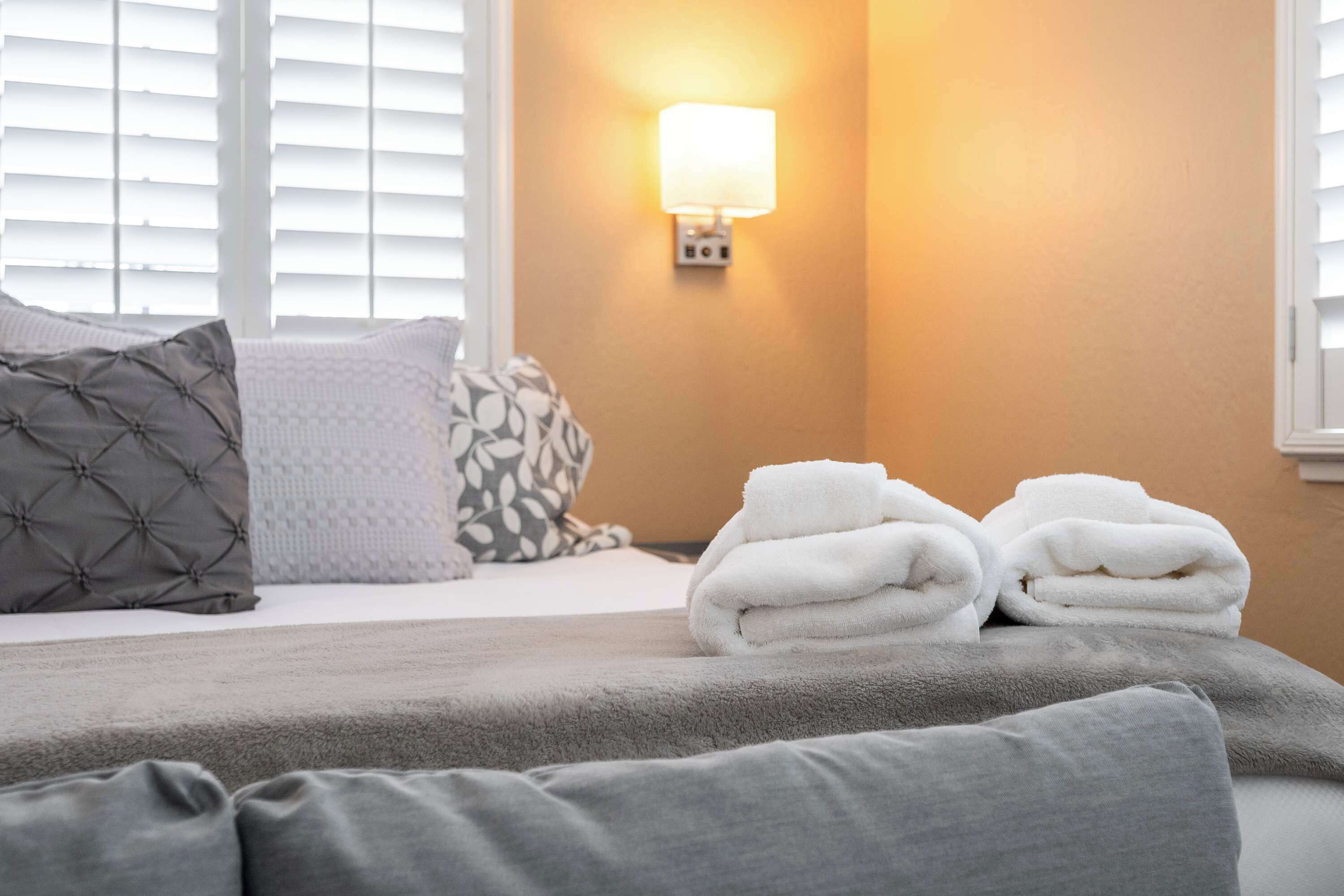 A neatly made bed with a grey blanket, two grey pillows, and a stack of white towels.