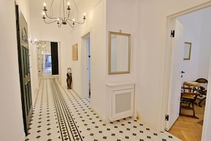 A hallway with patterned flooring, a chandelier, a mirror, and a door leading to a room with a chair.