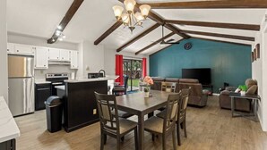 A modern kitchen with white cabinets, a black island, and stainless steel appliances. There is a dining area with a wooden table and chairs, and a living room with a blue accent wall and a TV. - Incredible Location | King Bed | Sleeps 14 (San Antonio)