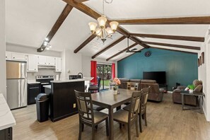 Eine moderne Küche mit weißen Schränken, einer schwarzen Kochinsel und Edelstahlgeräten. Es gibt einen Essbereich mit einem Holztisch und Stühlen, sowie ein Wohnzimmer mit blauer Akzentwand und einem Fernseher.