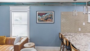 A modern kitchen with a granite countertop, a dining area with a wooden table and chairs, a blue accent wall, and a framed cityscape poster. - The Stafford Cottage-Close to The Pearl & Downtown (San Antonio)
