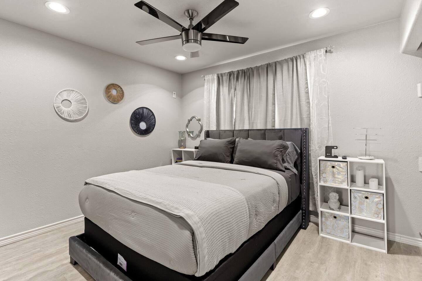 Ein Schlafzimmer mit einem Bett, einem Deckenventilator, einem Nachttisch und einer kleinen, weißen Regaleinheit.