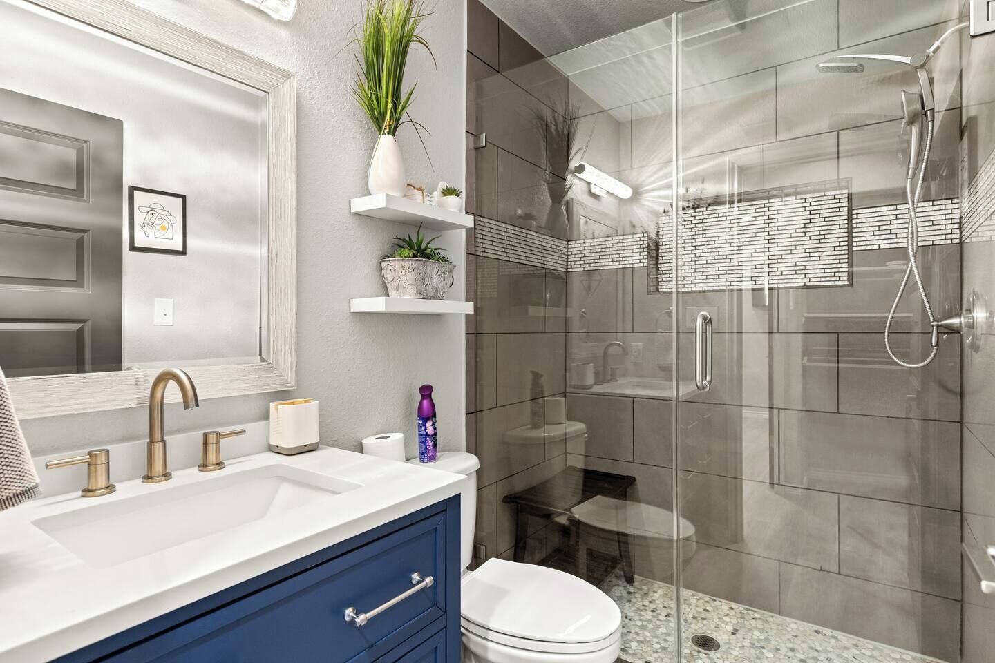 Un bagno moderno con doccia vetrata, lavandino bianco con dettagli dorati e un mobiletto blu.