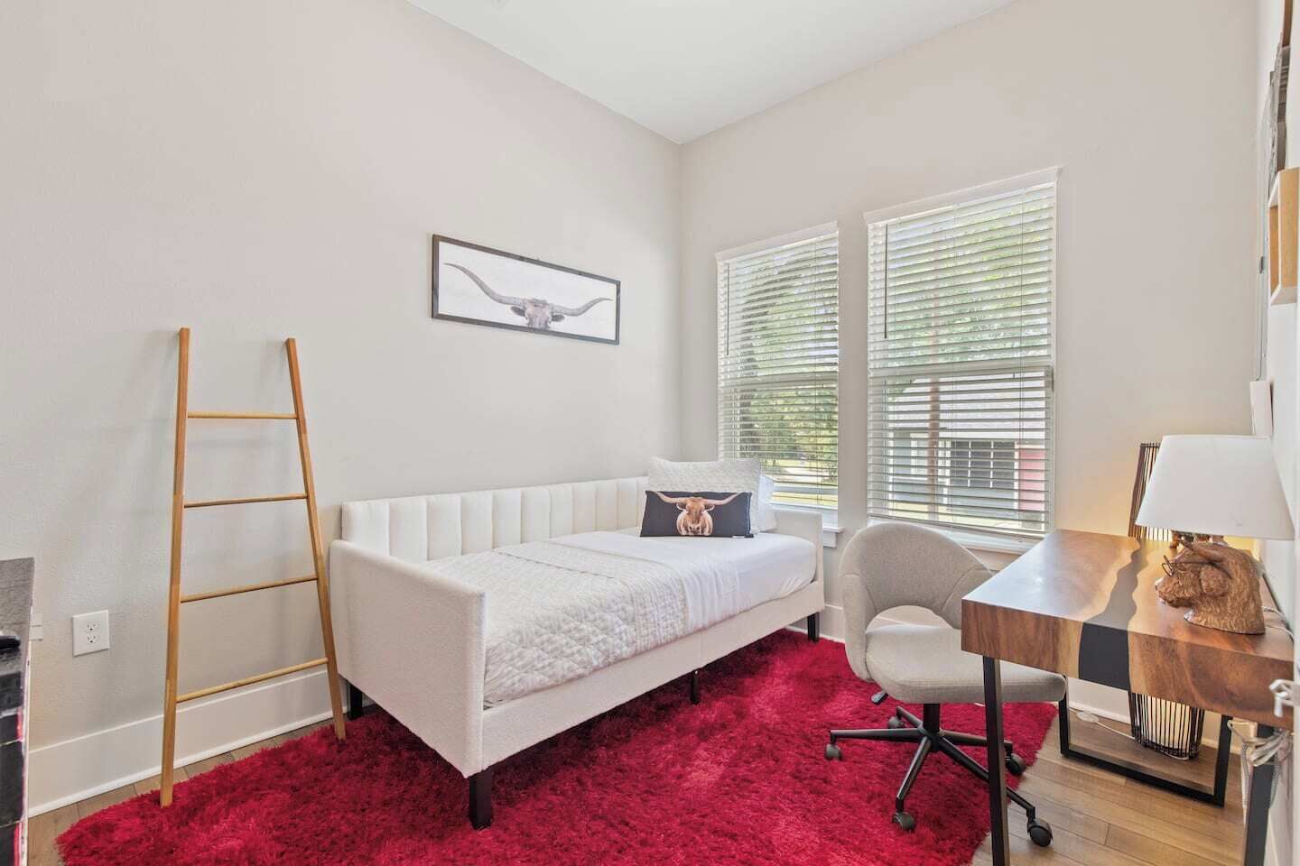 A room with a white sofa, a red carpet, a desk with a lamp, and a window with blinds.