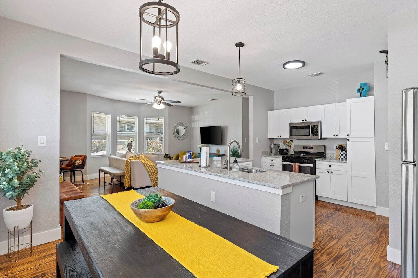 Une cuisine moderne avec des meubles blancs, un îlot central et un coin repas comprenant une table en bois et un chemin de table jaune.