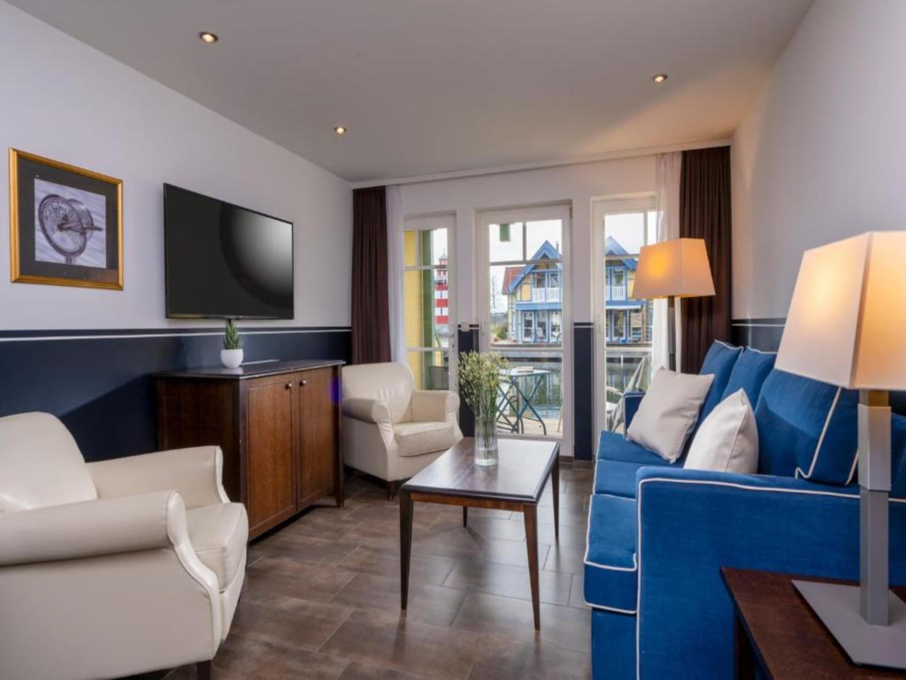 A modern living room with a sofa, armchair, TV, and a view of buildings through large windows.