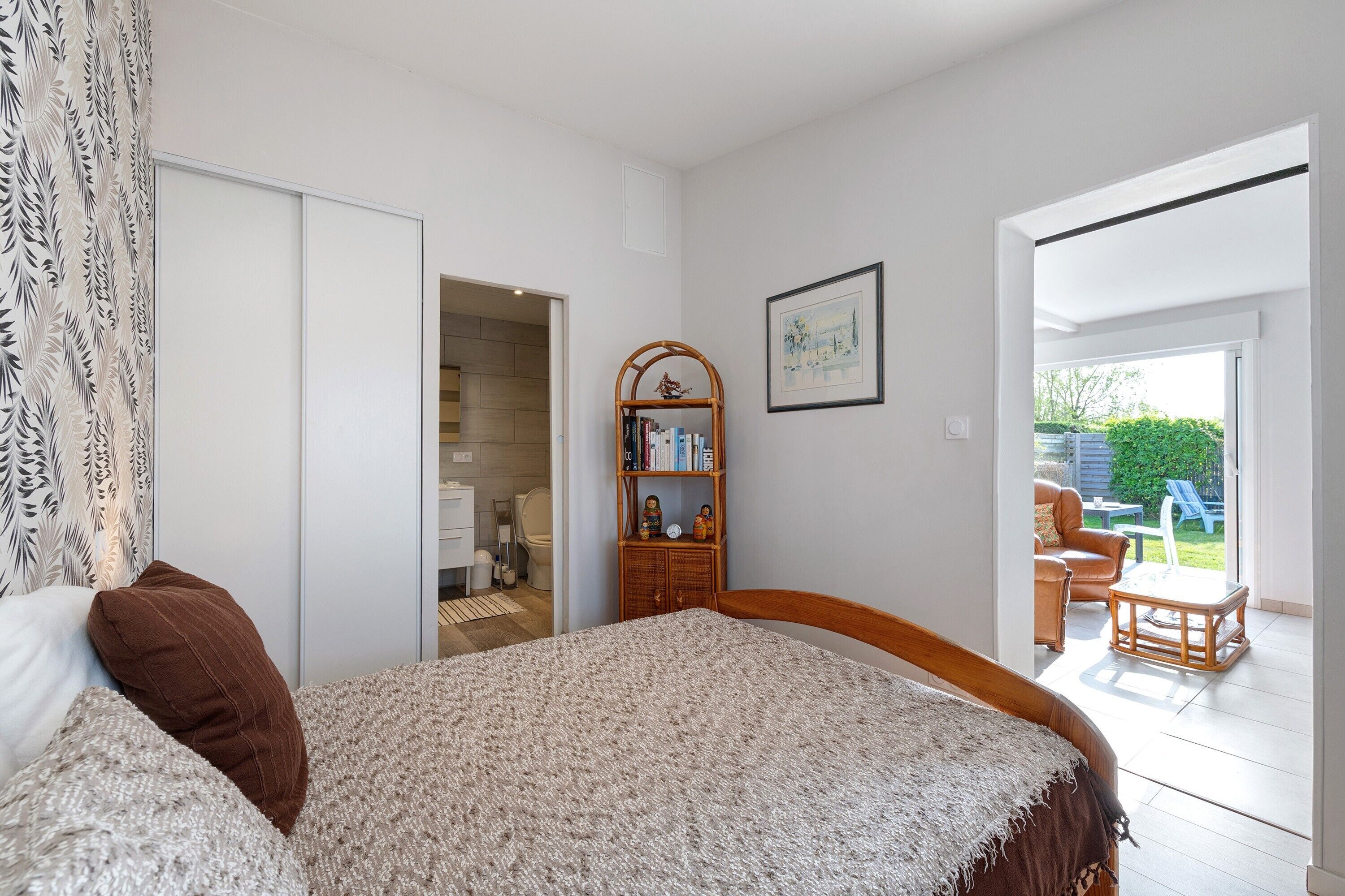 A bedroom with a bed, a wardrobe, a small wooden shelf with books, a framed picture, and a view of an outdoor patio.