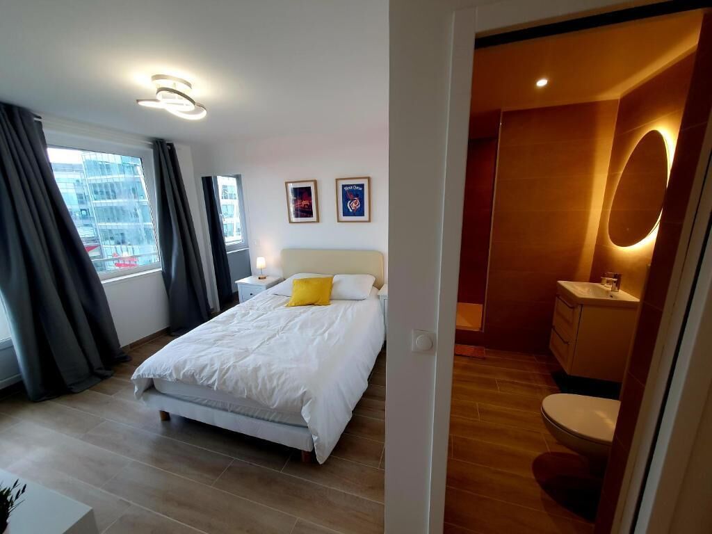 Ein modernes Hotelzimmer mit einem großen Bett, einem Nachttisch, einem Fenster mit Vorhängen sowie einem Badezimmer mit Waschbecken und Toilette.