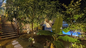 Ein Garten bei Nacht mit einer Steintreppe, einem kleinen Gebäude mit Strohdach und einem Poolbereich.