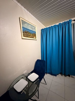 A small room with a single chair, a table, a painting on the wall, and a window with blue curtains.