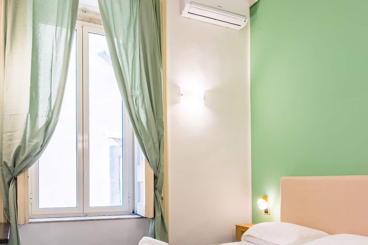 A room with a bed, green curtains, an air conditioning unit, a nightstand, and a wall-mounted light.