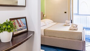Ein Hotelzimmer mit einem Bett, einem kleinen Tisch mit Pflanze und Bilderrahmen, einem Spiegel und Blick nach draußen.
