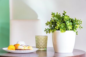 A white ceramic pot with green foliage, a plate with muffins and oranges, and a textured glass on a wooden table.