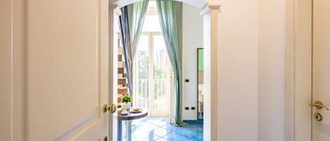 A hallway with a blue tiled floor, a white door with a number, and a staircase leading to a bright room with a window.