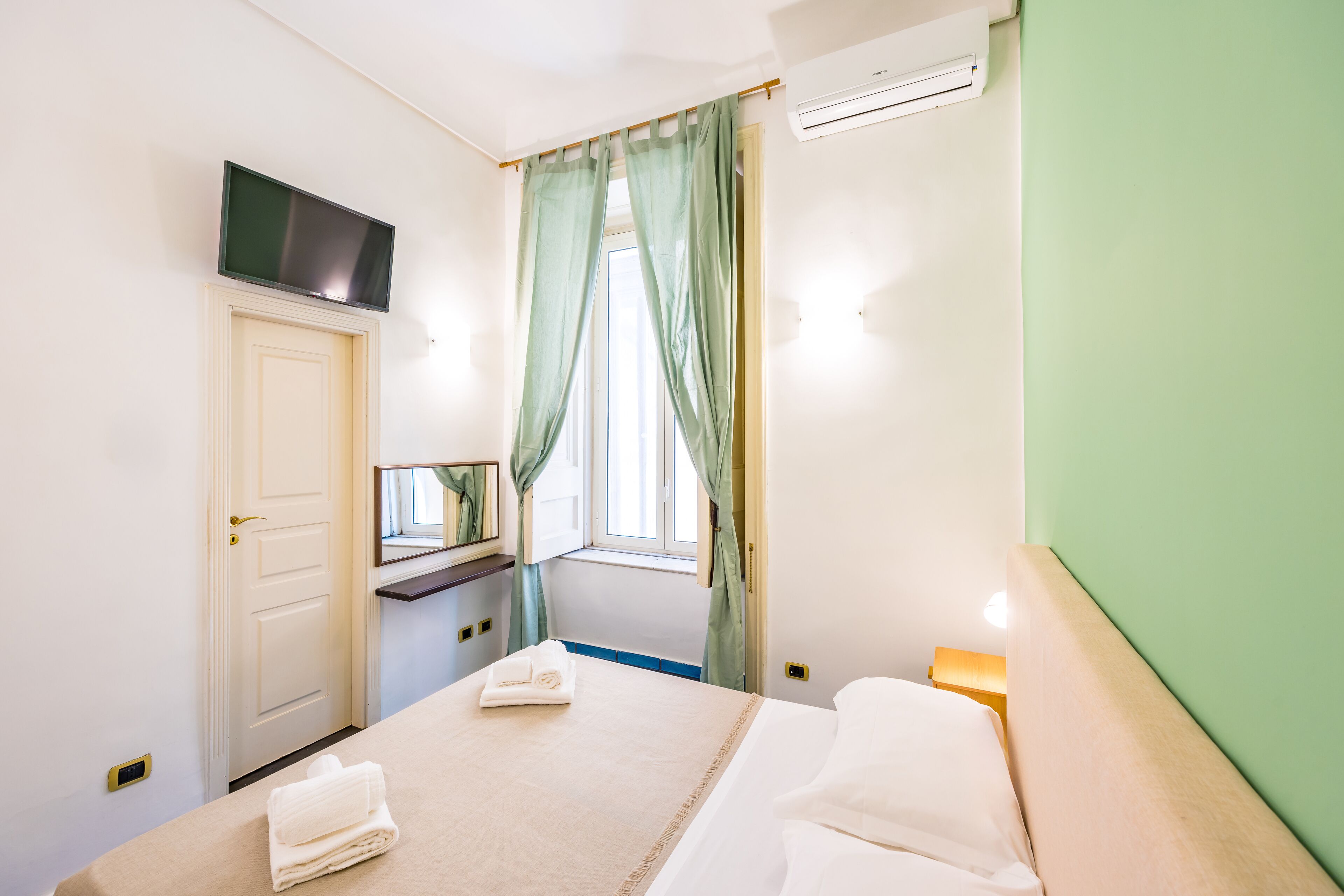A hotel room with a bed, a flat-screen TV mounted above the door, a window with curtains, and an air conditioning unit.