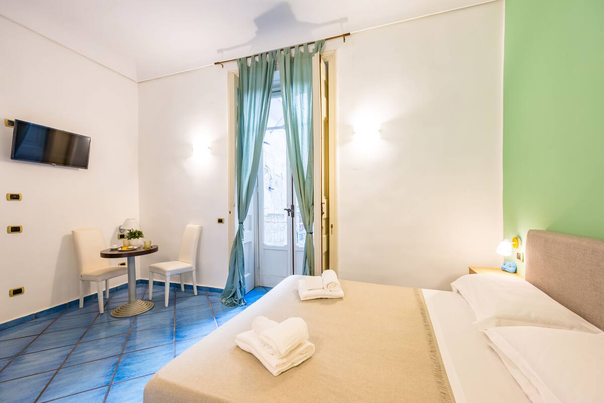 A hotel room with a bed, two chairs, a small table with a lamp, a TV mounted on the wall, and a window with curtains.