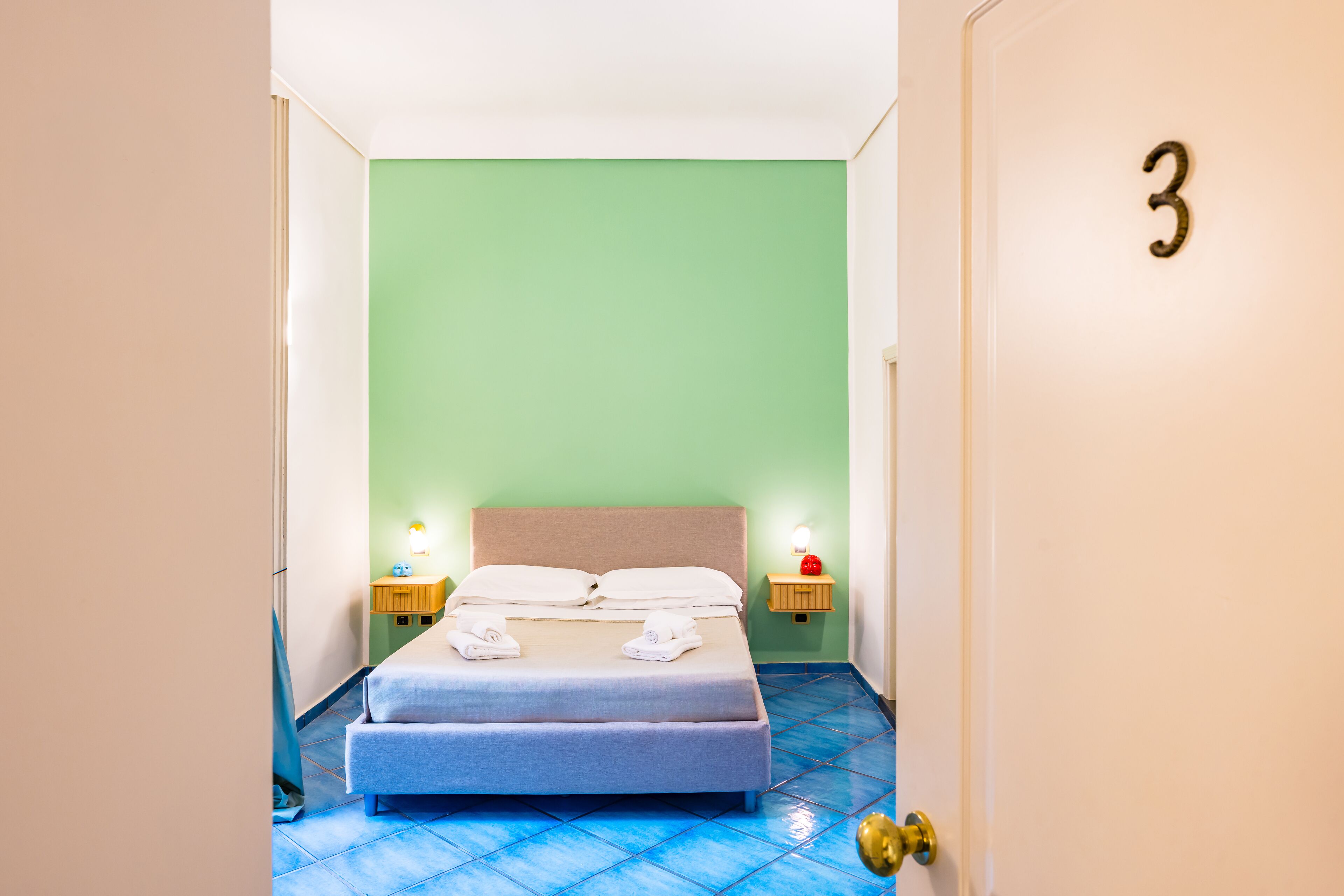 Habitació d'hotel amb un llit, dues taules de nit amb làmpades i una porta amb el número 3.