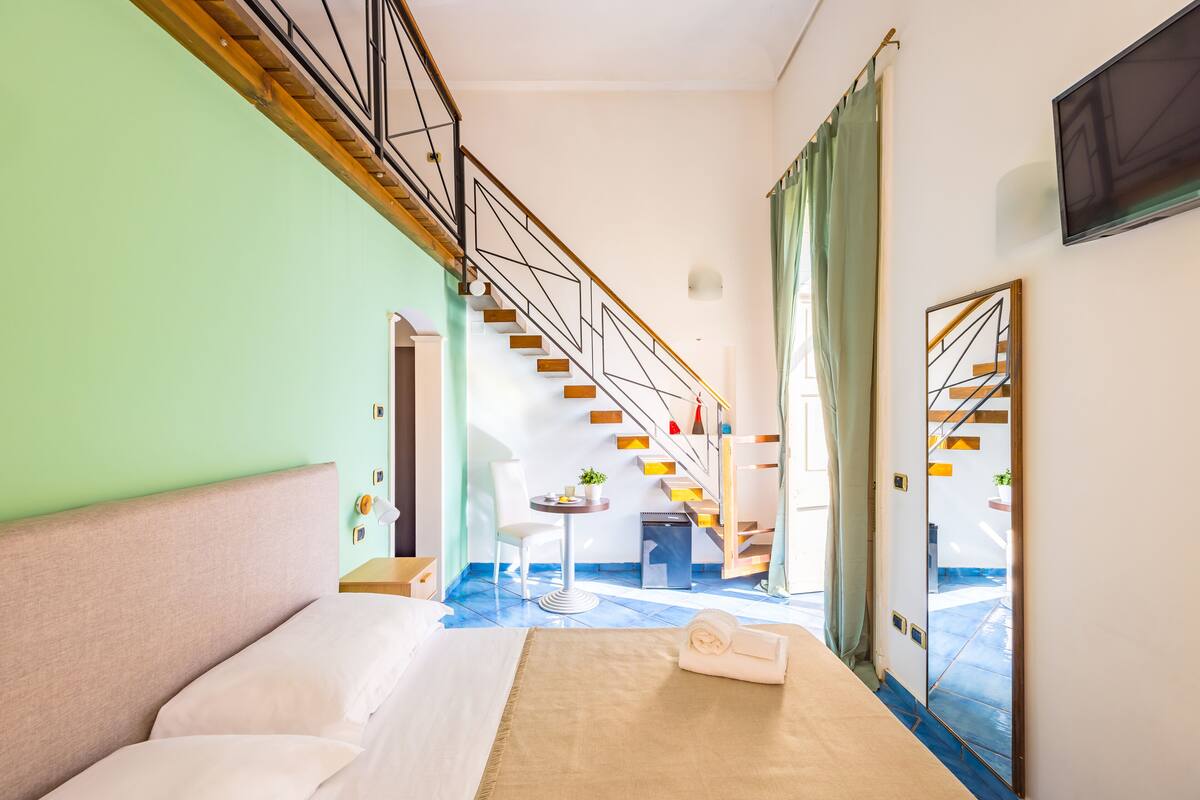 A hotel room with a bed, a small table with a plant, a chair, a staircase, and a view of the outside.