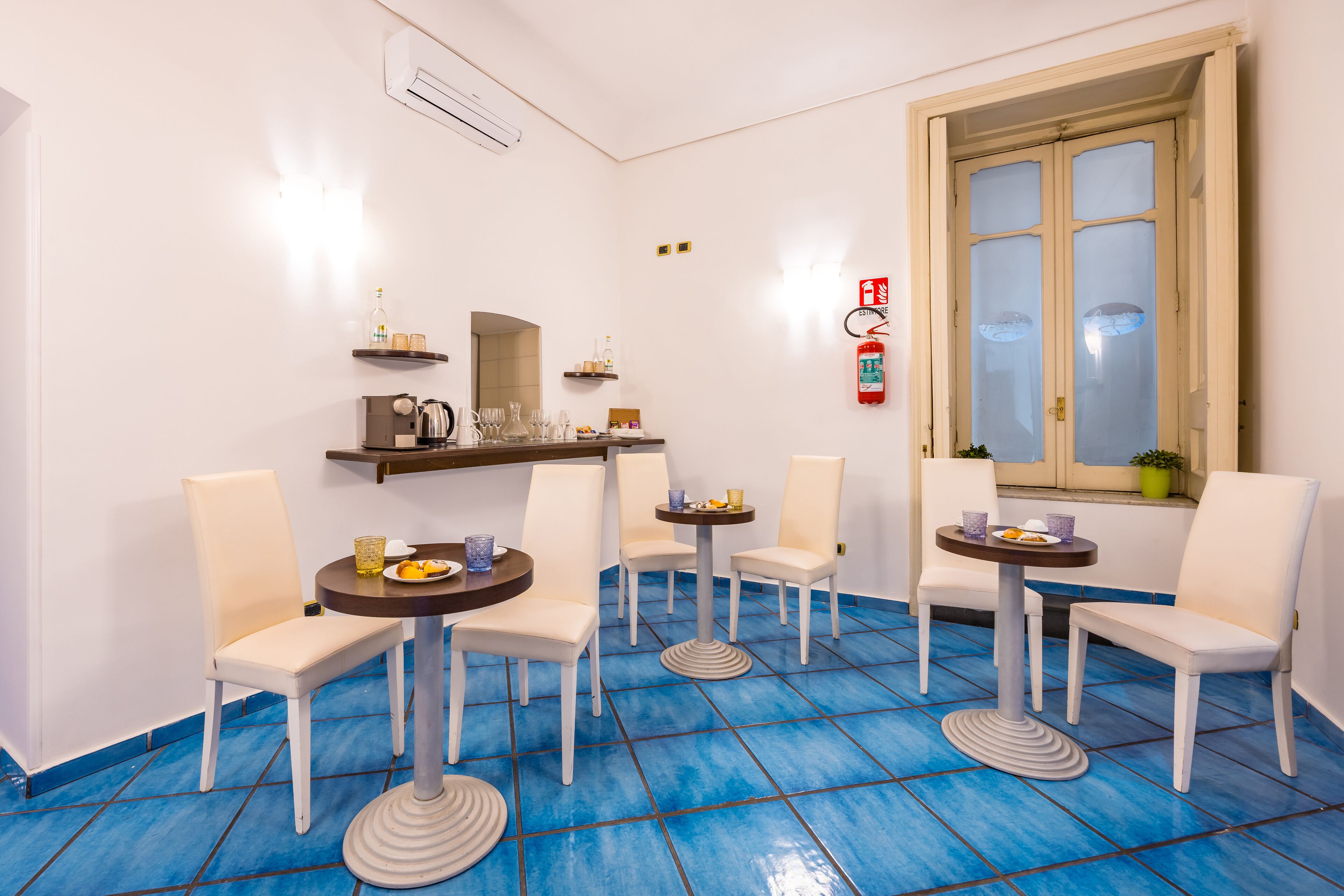 A dining area with white chairs and round tables, blue tiled floor, and a window with frosted glass.