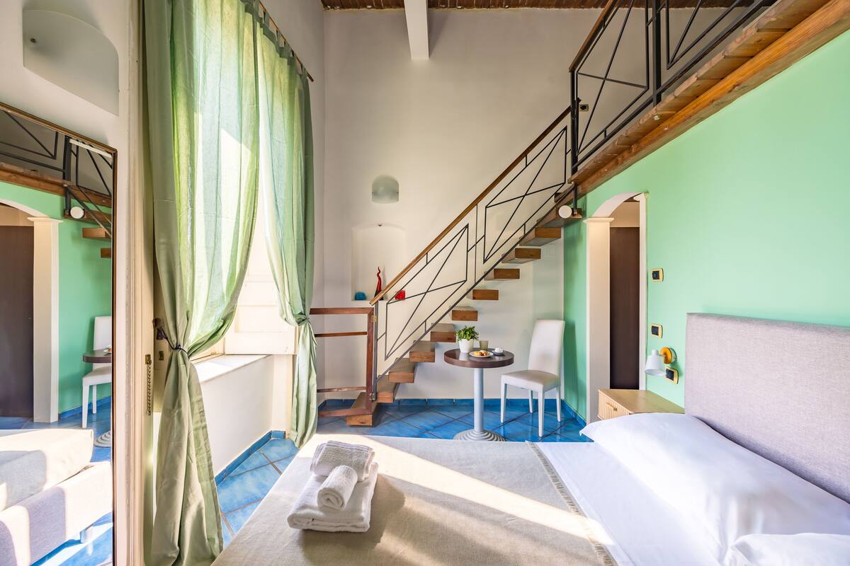 A hotel room with a bed, a small table, a chair, a staircase, and a view of the outside.
