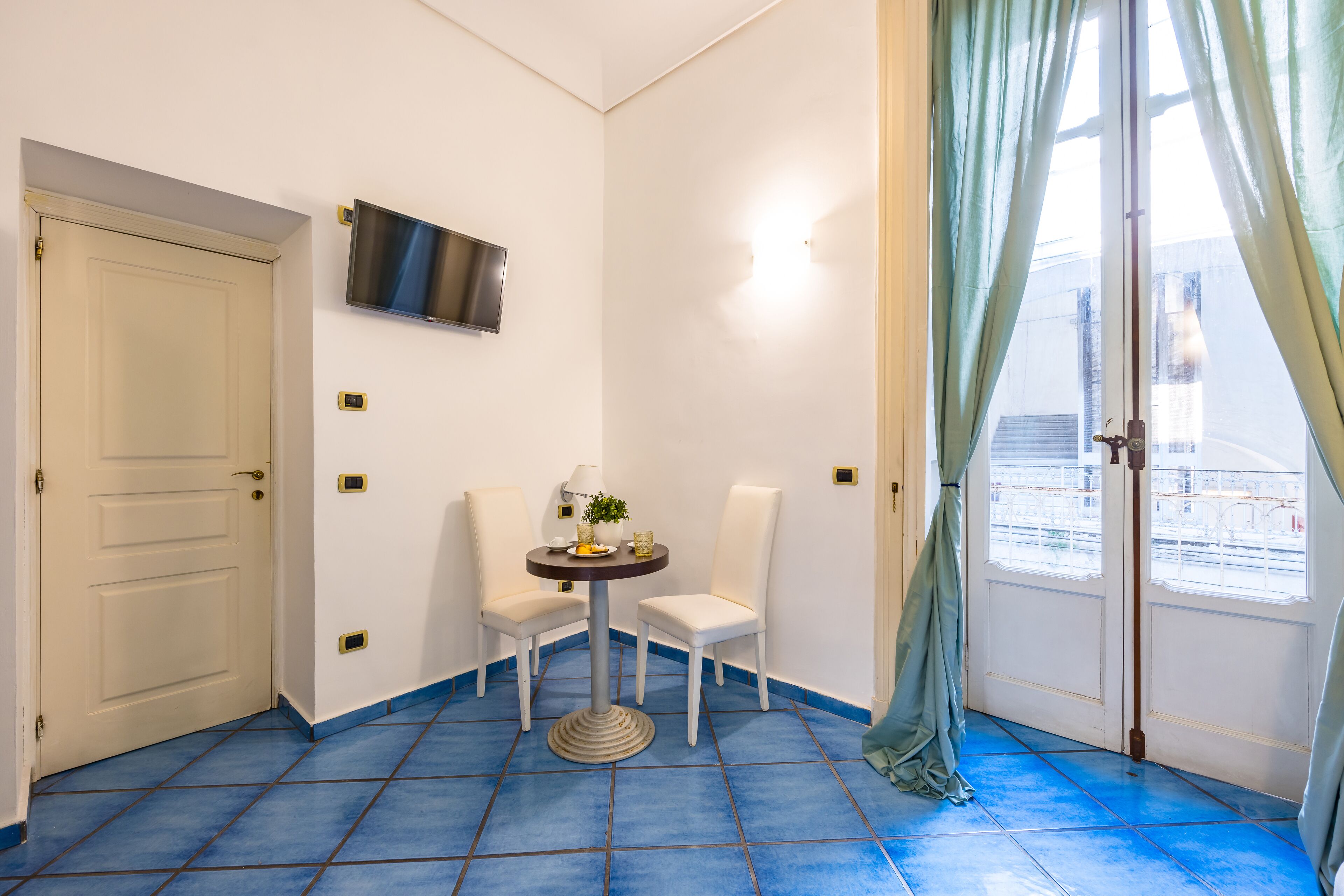 A room with a white door, a flat-screen TV mounted on the wall, a small round table with chairs, and a window with curtains.