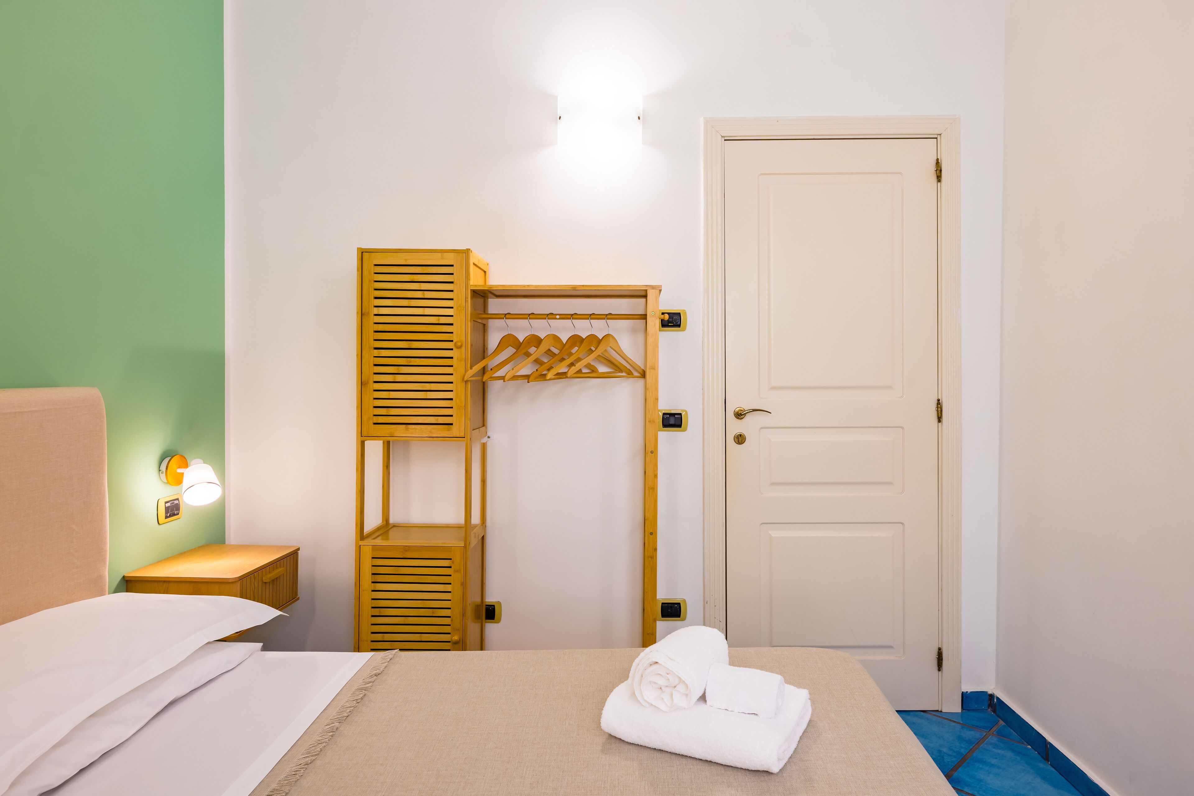 A hotel room with a bed, a wooden wardrobe, a hanging clothes rack, a door, and a nightstand with a lamp.