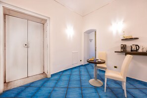 A room with a blue-tiled floor, a white door, a round table with a chair, and a shelf with a kettle and other items.