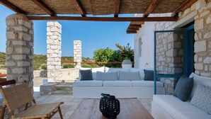 A stone-walled outdoor seating area with a wooden table, a woven chair, and a white sofa with patterned cushions. - Sand Key Villa 1 - A beautiful cycladic residence with 7 Bedrooms (Paros)