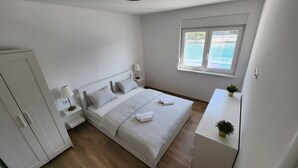 Een slaapkamer met een bed, nachtkastjes, een raam met uitzicht op zee, en een kleine plant op een nachtkastje.