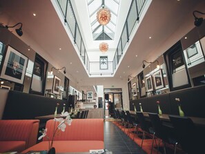 Un moderno vestíbulo de hotel con techos altos, un atrio central y un área de descanso con sofás naranja y sillas negras.