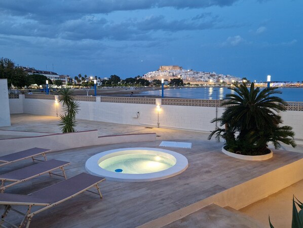 A rooftop pool area with lounge chairs, a hot tub, and a view of a coastal town at dusk.