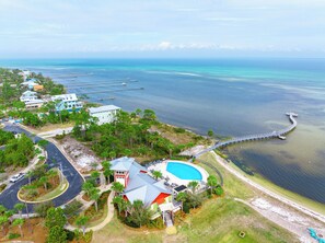An aerial view of a coastal property with a swimming pool, a pier extending into the water, and a road curving around the property.
