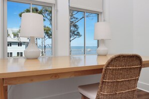 A wooden desk with two white lamps and a wicker chair in front of a window with a view of the ocean and buildings.
