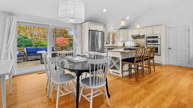 A modern kitchen with white cabinets, a stainless steel refrigerator, and a dining area with a glass table and chairs. - Cape Cod Lake Retreat on Angus Way RPMS169 (Centerville)