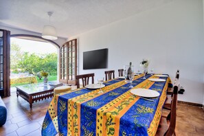A dining area with a long table covered in a patterned tablecloth, chairs, a television mounted on the wall, and a view of greenery outside. - Apartment in a villa with a private garden, 600 meters from the beaches (San Lorenzo al Mare)