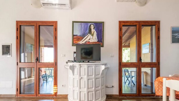 A room with wooden flooring, a white cabinet with a flat-screen TV, and two glass doors with wooden frames. - Delightful and Spacious Villa in Capo Comino (Siniscola)