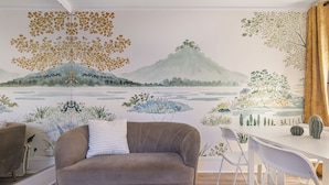 Een kamer met een bank, een eettafel en stoelen. Op de muur bevindt zich een muurschildering van een landschap met bomen en bergen.