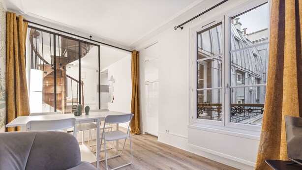 Een moderne woonkamer met een bank, een eettafel met stoelen en een groot raam met uitzicht op een gebouw.