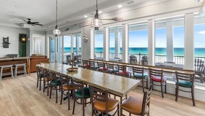 A spacious dining area with a long table and chairs, large windows with a view of the ocean, and a ceiling fan. - Newly built | Luxury Gulf-front Home | Private Pool | Elevator | Game Room (Pensacola Beach)