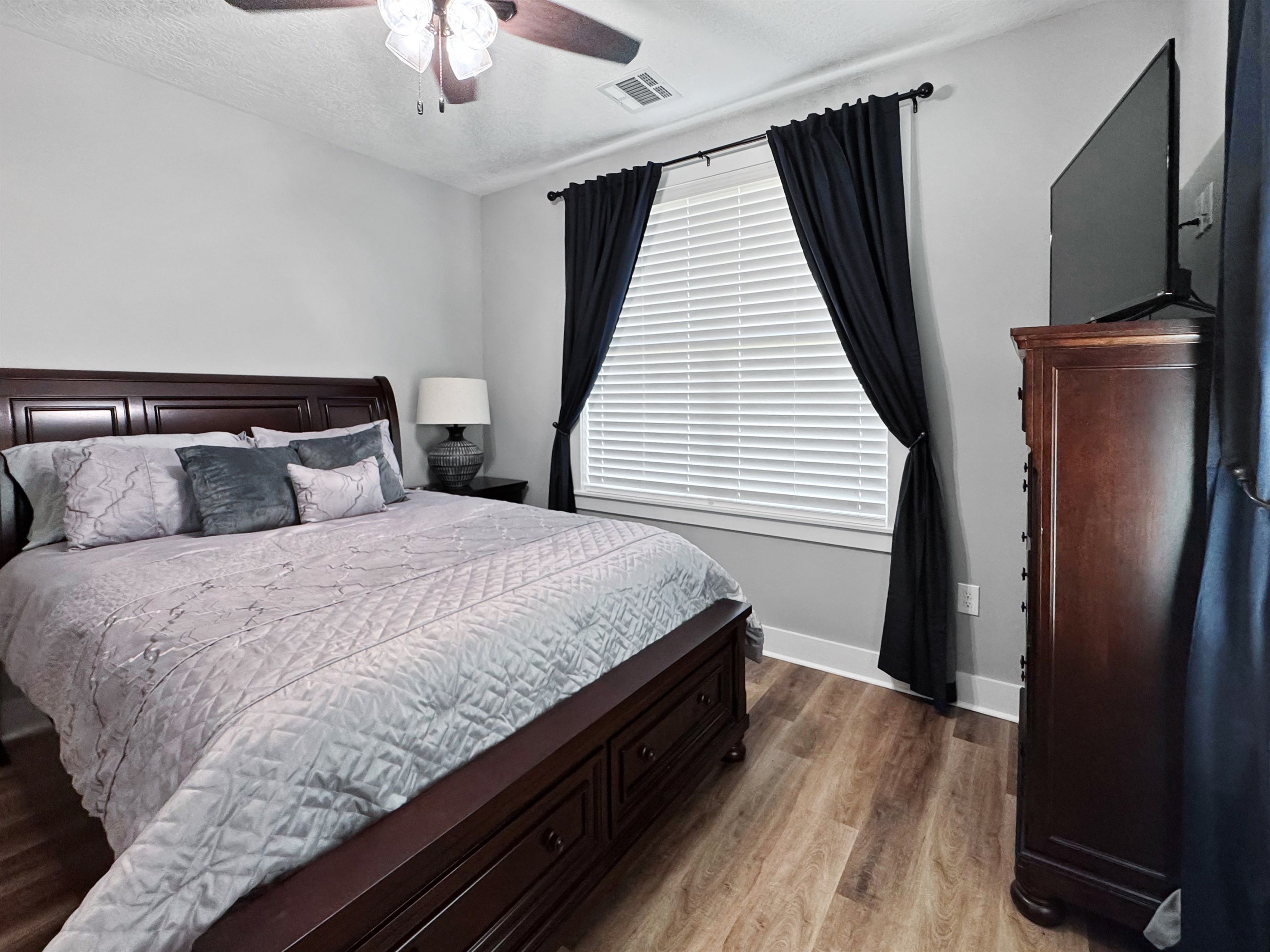Un dormitorio con cama, mesita de noche, lámpara, ventilador de techo, ventana con cortinas y televisor en la pared.