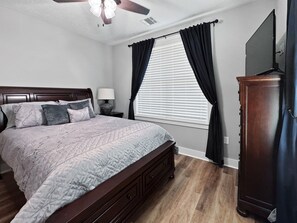A bedroom with a bed, a nightstand, a lamp, a ceiling fan, a window with curtains, and a TV on the wall.
