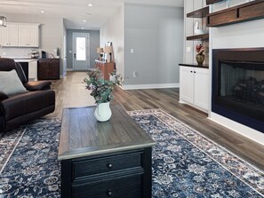 A living room with a dark brown sofa, a white coffee table, a blue patterned rug, a fireplace, and a kitchen in the background.