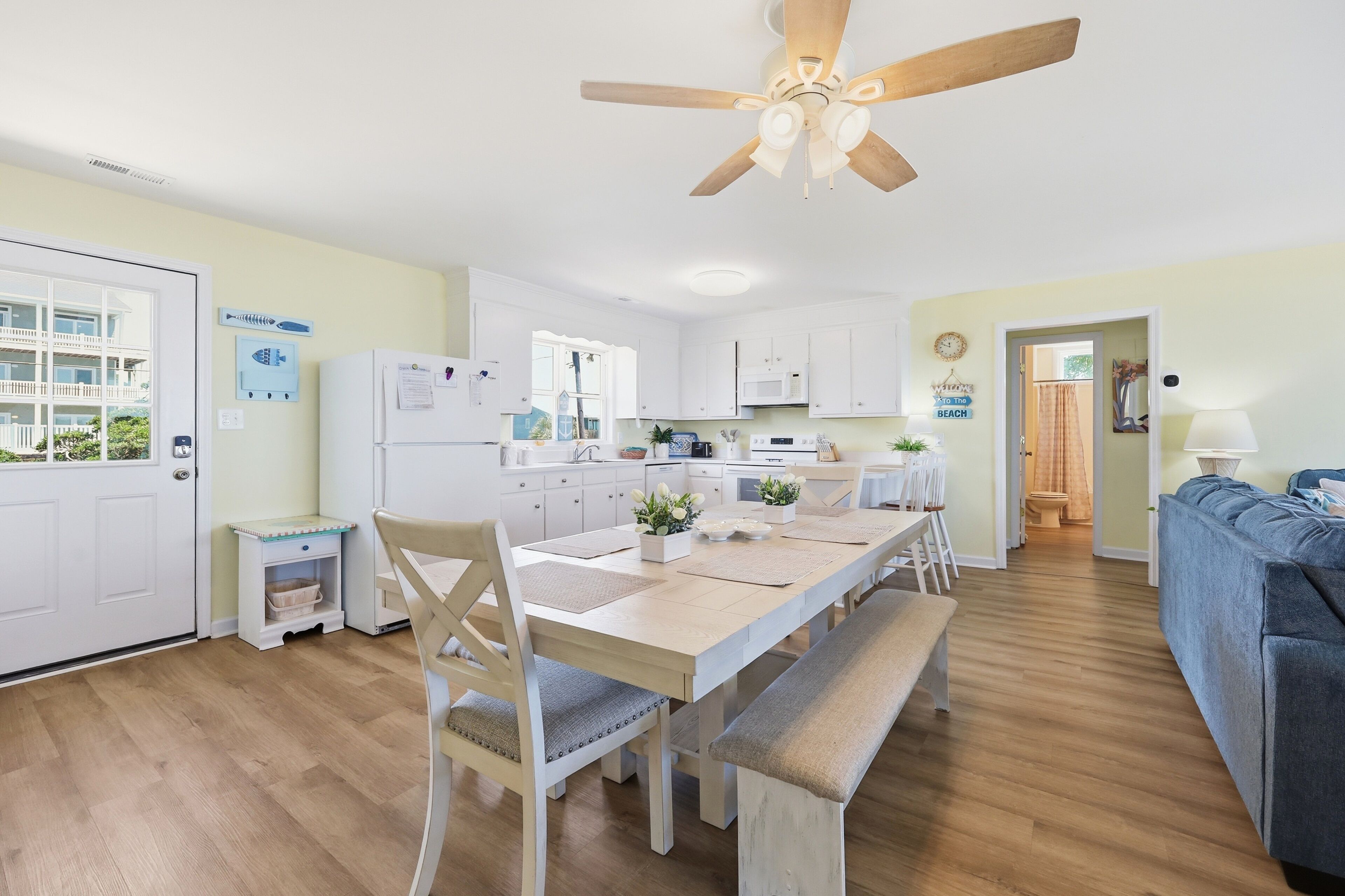 Une cuisine moderne avec des meubles blancs, un coin repas avec une table et des chaises en bois, un ventilateur de plafond et une vue sur un balcon.