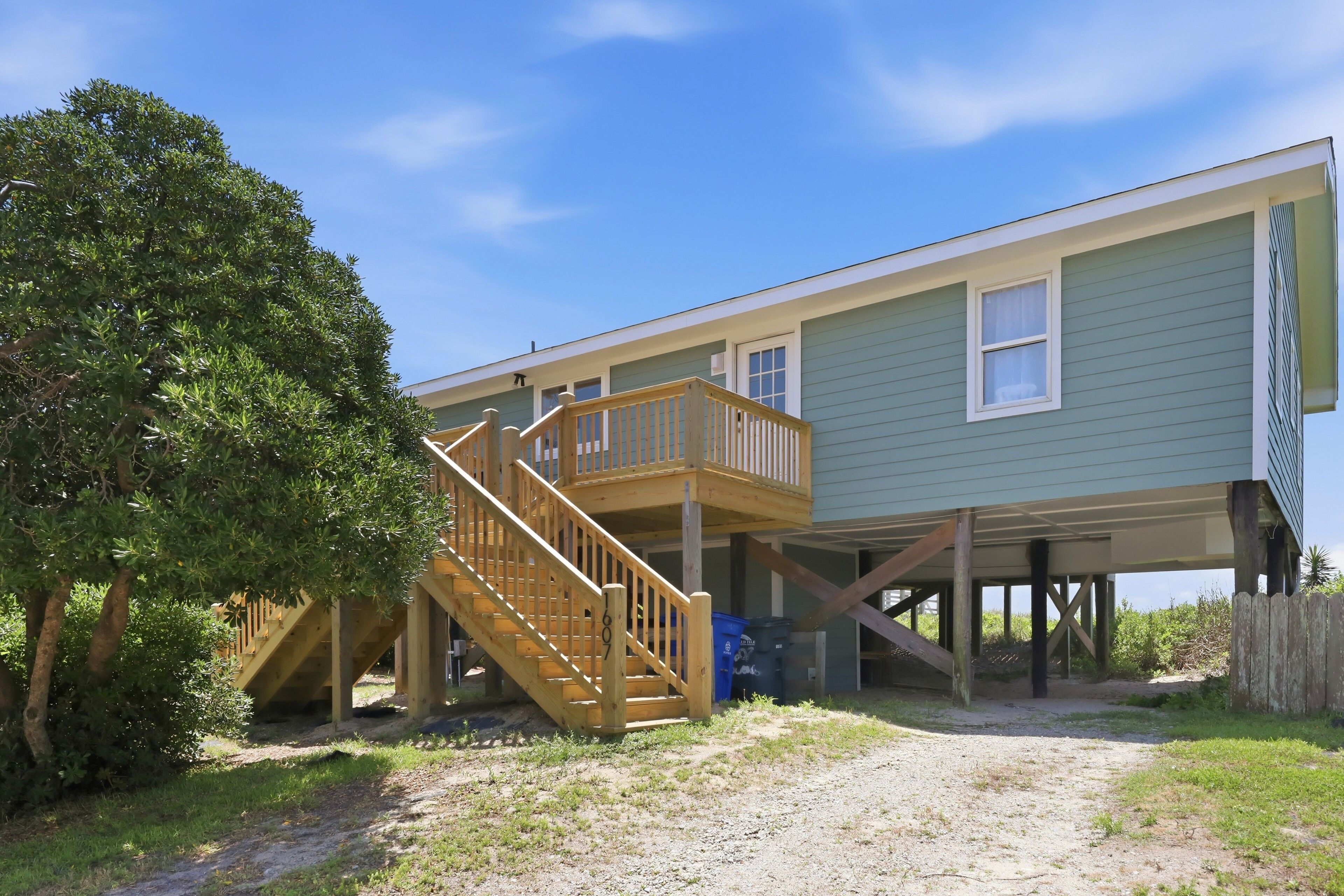 A house with a wooden staircase leading to a deck, a covered porch, and a sandy ground area.