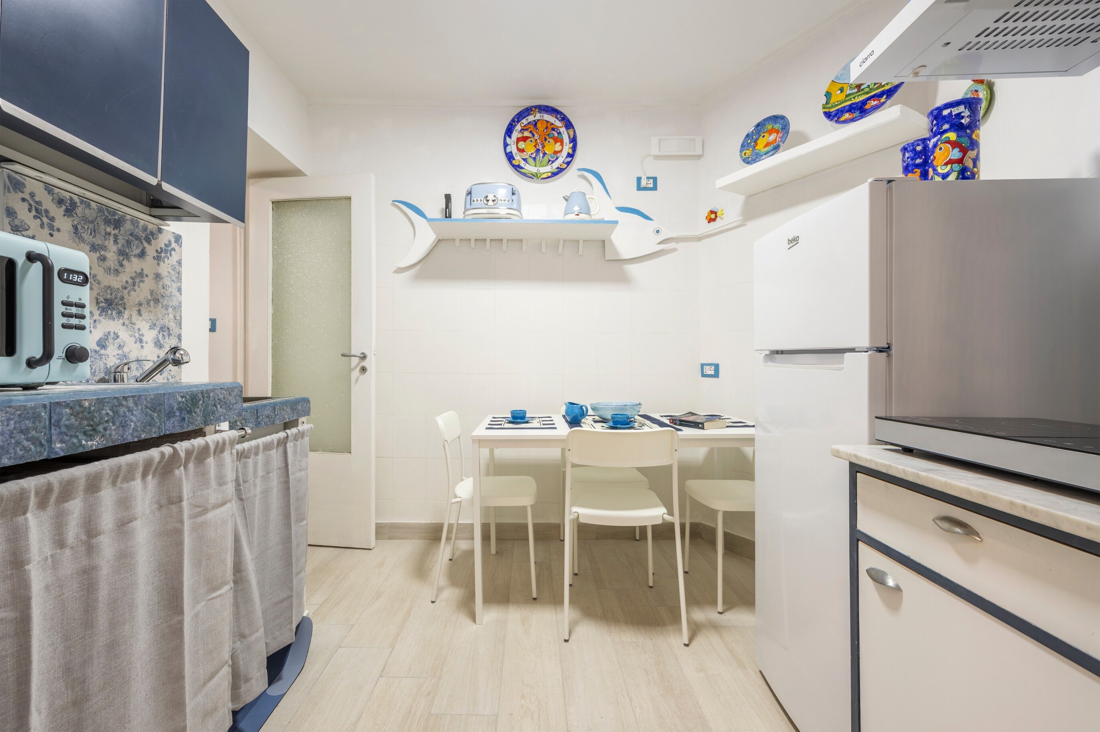 A compact kitchen with a microwave, stove, and refrigerator. There is a dining table with chairs and a small shelf above it.